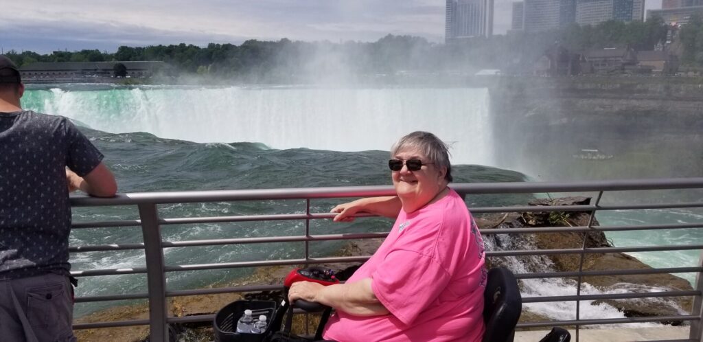 A woman with salt and pepper hair sits on an electric scooter in front of a waterfall.